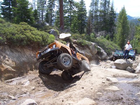1998 Ford Ranger - An orange off-road vehicle navigates a challenging rocky terrain, with other vehicles and spectators observing the action.