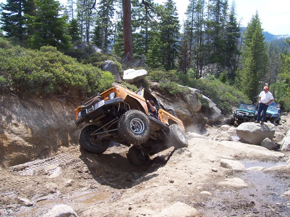 Orange off-road vehicle climbing a rocky trail, with other vehicles and people in the background
