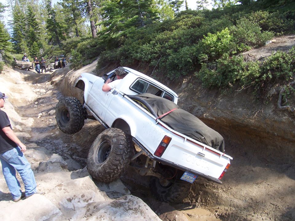 White pickup truck with large tires navigating a rocky off-road trail. Another vehicle is visible further up the trail