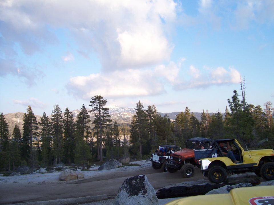 Yellow Jeep Wrangler and other off-road vehicles on a dirt path with trees and mountains in the background
