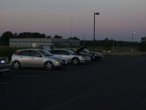 1998 Ford Taurus - Several cars are parked in a lot as the sky darkens, with one sedan's hood open.