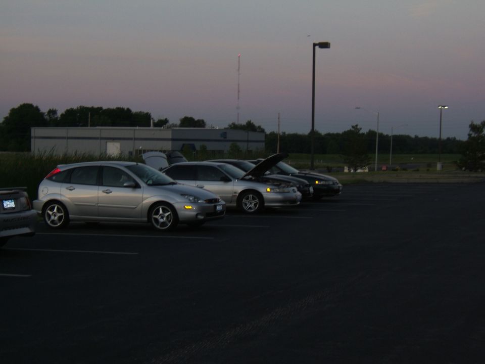 silver Ford Focus hatchback, silver sedan with hood up, dark sedan, and part of a gray car in a parking lot at dusk
