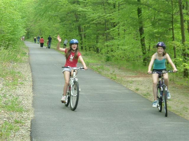 Girl in red shirt waves while riding a bike, another girl in blue top rides behind her on a wooded path