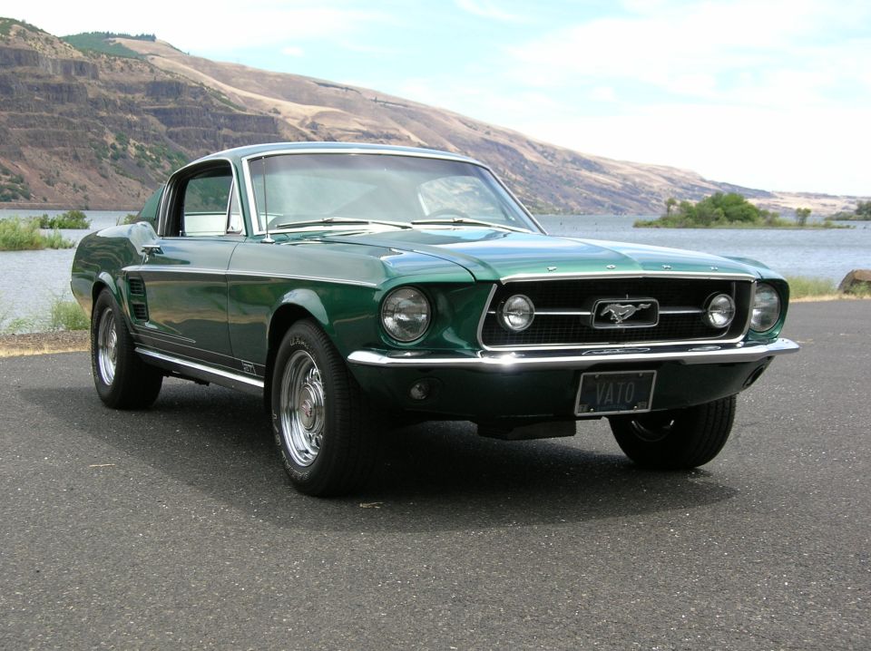 Dark green Ford Mustang fastback, 3/4 front view, parked by a river with cliffs behind