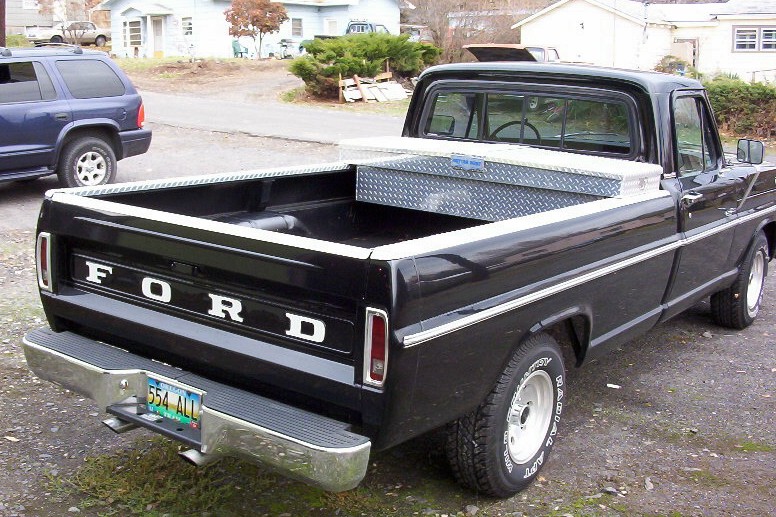 Black Ford pickup truck parked outside with a toolbox in the bed. Blue SUV in background