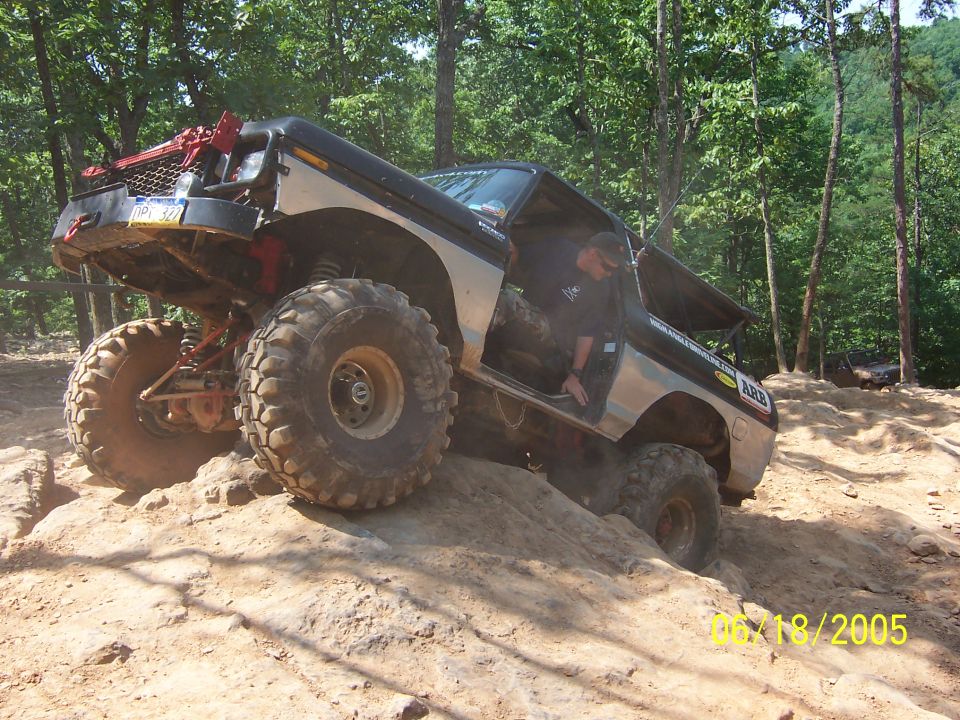 Modified off-road SUV with large tires climbing a steep dirt incline. Driver is visible in the open door