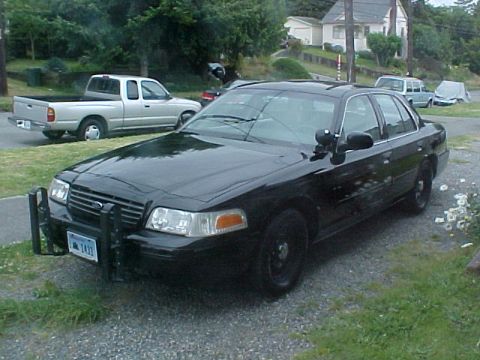 1999 Ford Crown Victoria - A black police car is parked on a gravel driveway, with a silver pickup truck in the background.