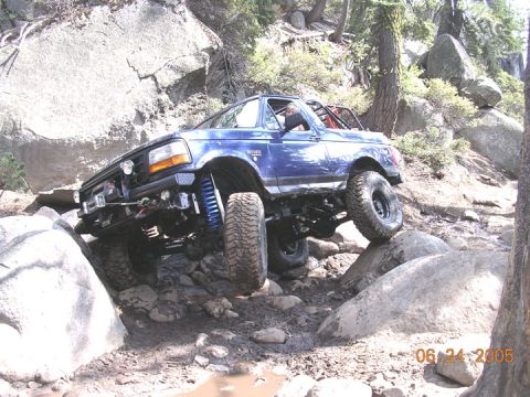 1994 Ford Bronco - A blue 1994 Ford Bronco tackles a challenging off-road course with large rocks and mud.