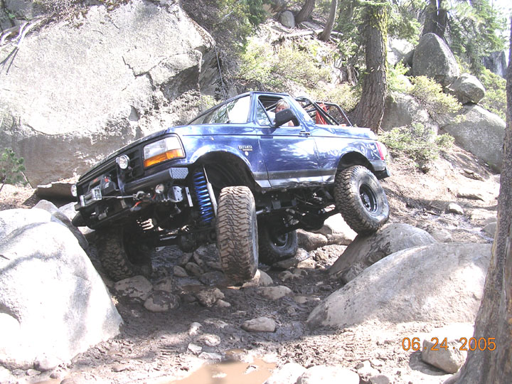 Blue 1994 Ford Bronco navigating rocky terrain, front-left tire elevated