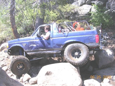 1994 Ford Bronco - A blue Ford Bronco is shown off-roading over large rocks on a sunny day.