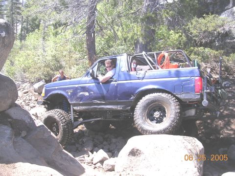 1994 Ford Bronco - A blue 1994 Ford Bronco is shown off-roading over large rocks on a sunny day.