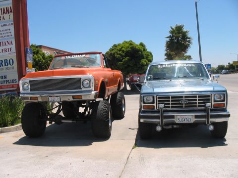 1986 Ford Bronco - Two lifted trucks, one orange and one light blue, are parked side-by-side on a sunny day.