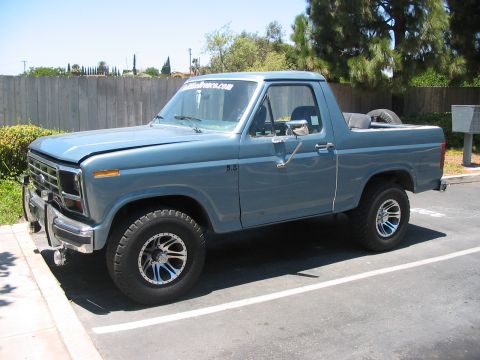 1986 Ford Bronco - This blue Ford Bronco is parked in a lot, viewed from the front three-quarter angle. The hood is open, and a spare tire is visible in the c…