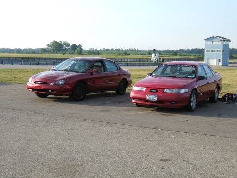 1998 Ford Taurus - Two red 1998 Ford Tauruses are parked side-by-side on a racetrack, with a blue building and tower in the background.