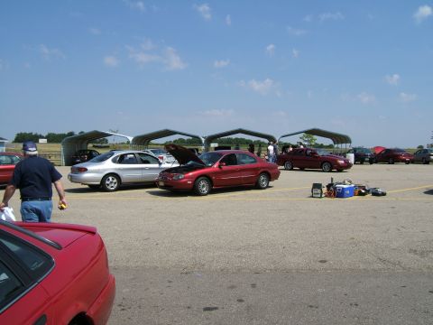 1998 Ford Taurus - A red 1998 Ford Taurus sits with its hood open at an outdoor gathering, surrounded by other vehicles.