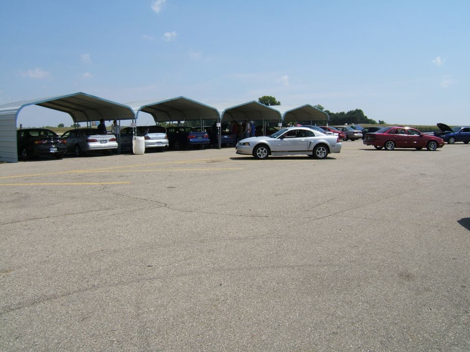 Silver coupe parked on asphalt, side view. Multiple cars under canopies in background