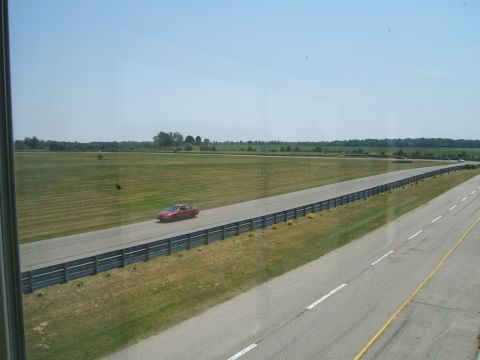 1998 Ford Taurus - A red sedan drives along a paved track bordered by a metal fence and grassy fields under a clear blue sky.