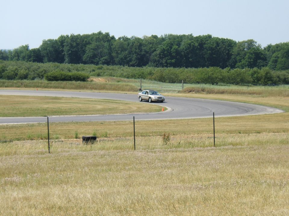 Silver sedan driving on a curving asphalt race track with grassy infield and trees in background