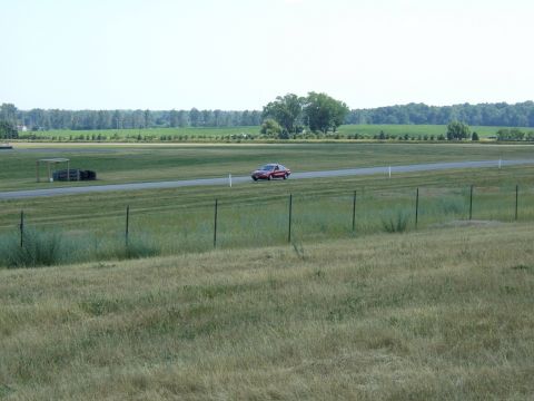 1998 Ford Taurus - A red sedan drives down a paved track surrounded by grassy fields under a clear sky.