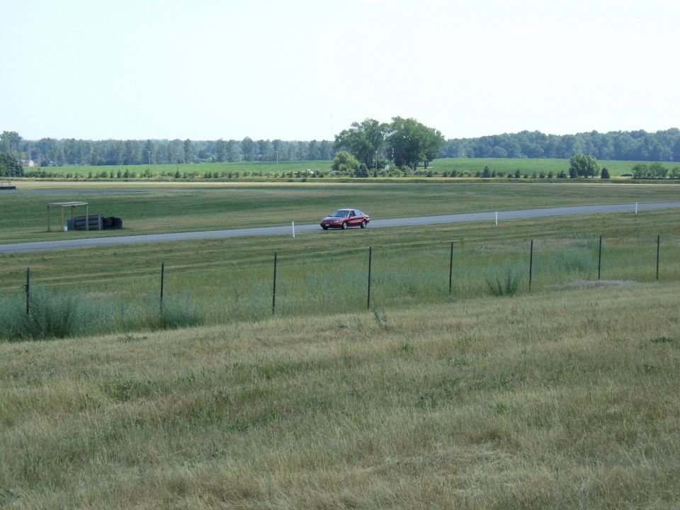 Red sedan driving on a racetrack with fields and trees in the background