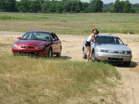 1998 Ford Taurus - Two Ford Taurus sedans are parked on a dirt road in a grassy field, with a woman interacting with one of the vehicles.