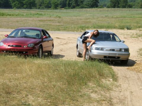 1998 Ford Taurus - A silver 1998 Ford Taurus is parked next to a red Taurus, with a woman posing on the hood of the silver car.