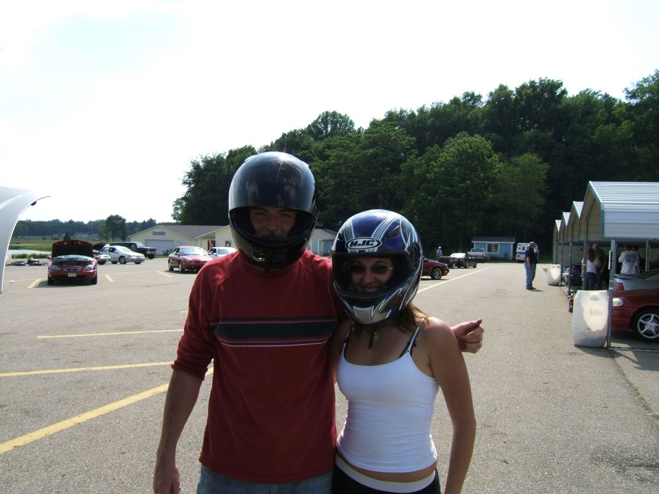 Two people in helmets at a track, with cars in the background