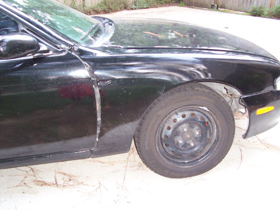 Front corner of a black coupe with bare steel wheel and rust-damaged fender