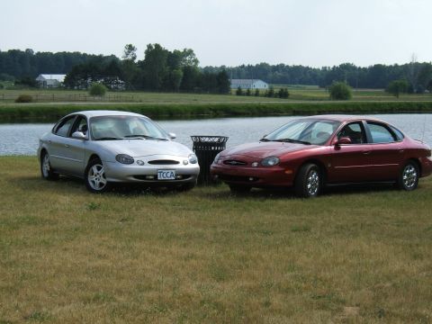 1998 Ford Taurus - Two Ford Taurus sedans, a silver 1998 model and a red one, are parked on a grassy area next to a lake.