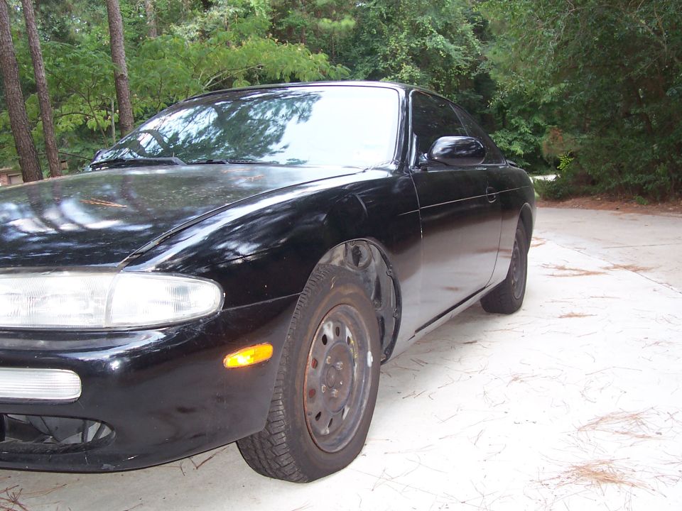 Black coupe on bare steel wheels parked on a concrete pad in a wooded yard, 3/4 front view