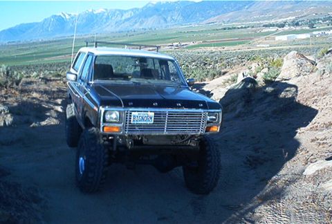 1986 Ford Bronco - A black Ford Bronco with a white roof is parked on a dirt road with mountains in the background.
