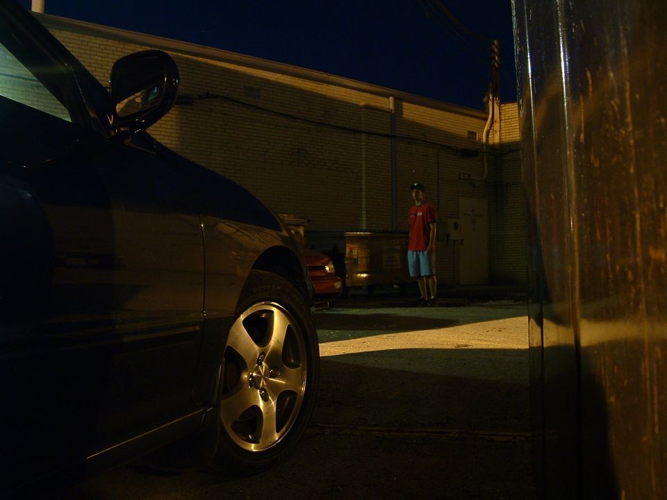 Dark colored car side view, parked in an alley at night. A person stands in the background near dumpsters