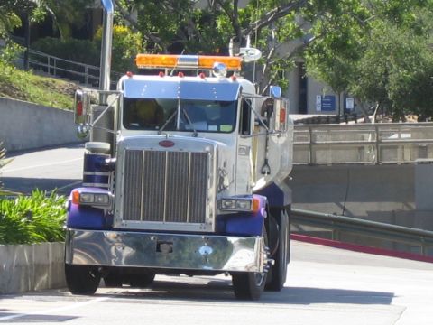 1990 Ford Bronco - Front view of a purple and white Peterbilt water truck with a chrome grille and amber light bar descending a paved ramp.