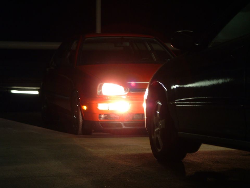 Red Volkswagen Golf Mk3 with headlights on, parked behind a dark car