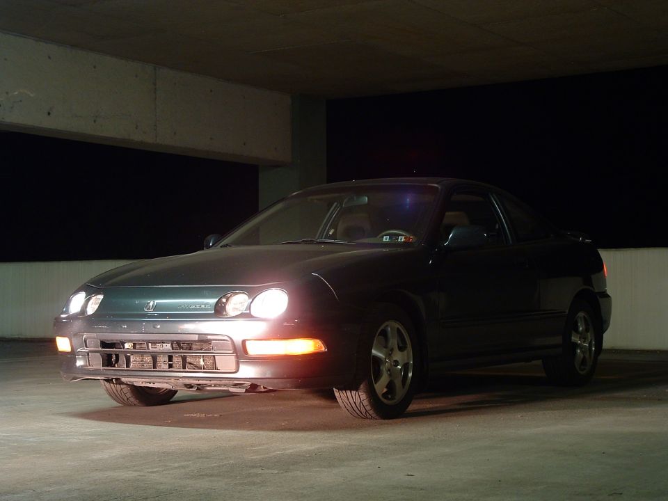 Dark coupe with headlights on, parked in a concrete structure. The badge on the front grille is visible