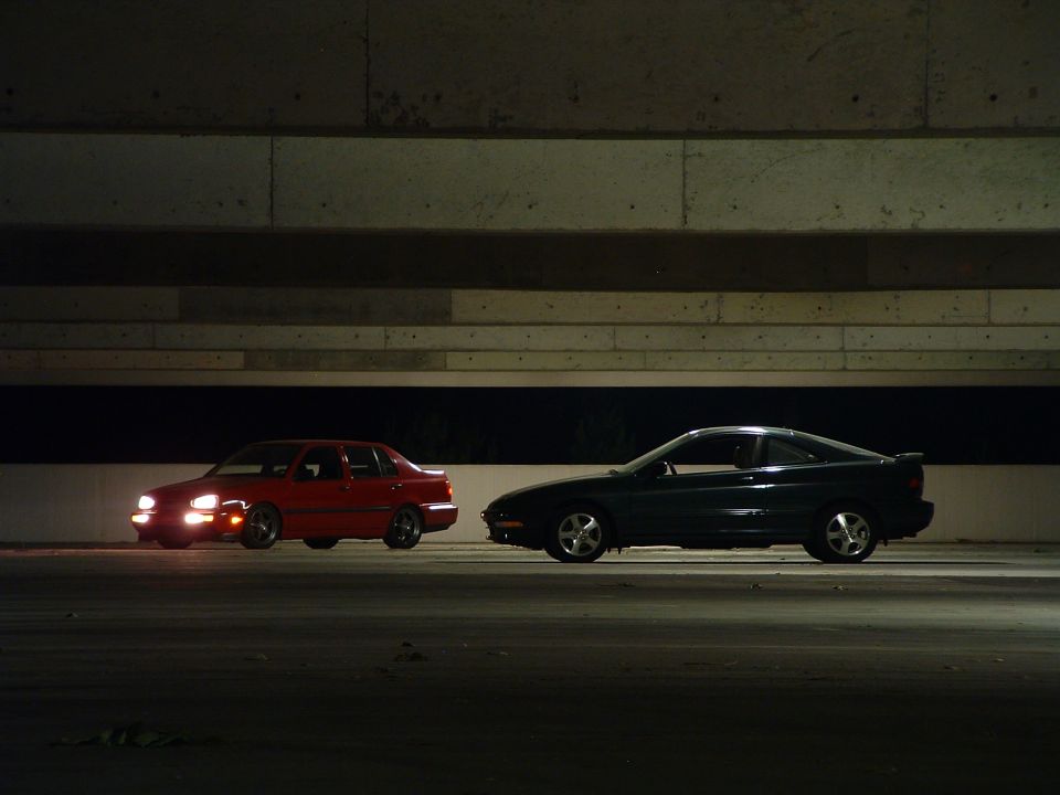 Red 1994 Volkswagen Jetta sedan and a dark coupe parked side-by-side under a concrete overpass at night