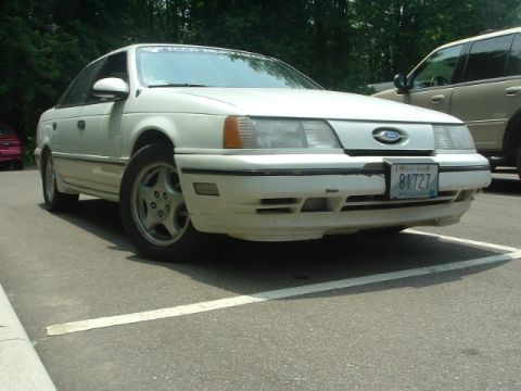 1993 Ford Taurus - A white sedan is parked in a lot, showing its front end and license plate. Another vehicle is partially visible to the right.