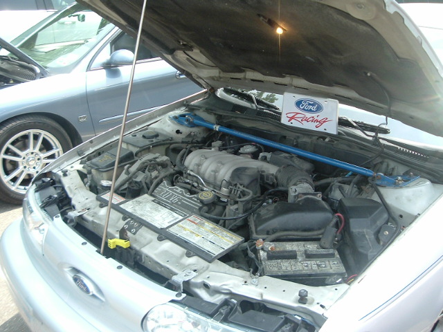 Engine bay of a 1993 Ford Taurus with hood open, showing the V6 engine and a Ford Racing sign. Another car is visible to the left