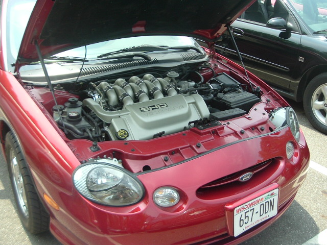 Red Ford Taurus SHO with hood open, showing V8 engine. Black car parked behind