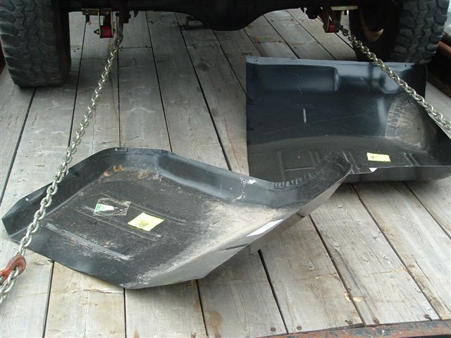 Two black metal floor pans, one curved and one flat, resting on wooden planks. Chains are attached to the underside of a vehicle above