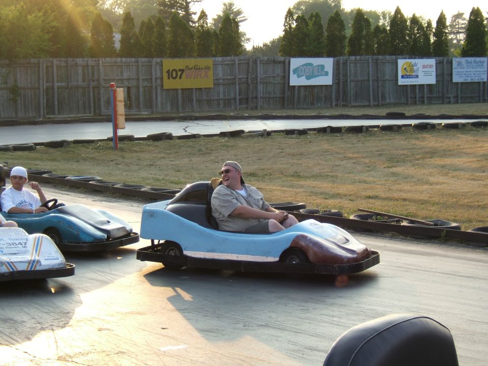 Guy in light blue go-kart smiling, another kid in blue go-kart behind him. Go-kart track with signs in background