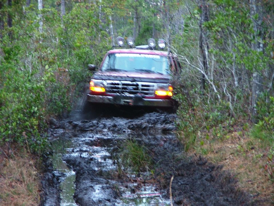 Muddy dark red Ford Bronco with roof lights driving toward camera on a rutted mud trail in the woods