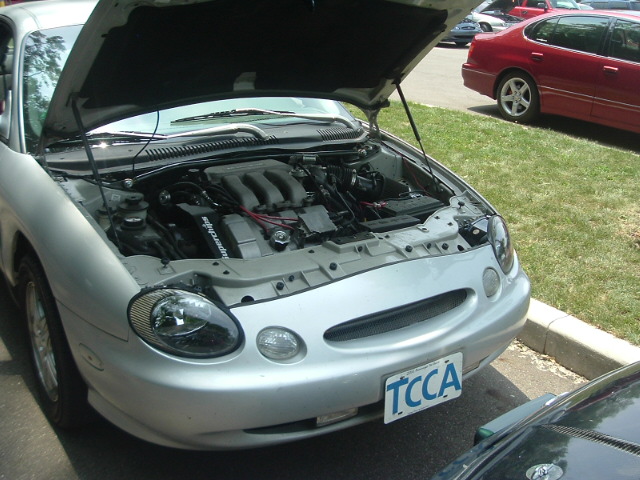 Silver Ford Taurus with hood open, revealing the engine. License plate reads TCCA. Another red car is parked nearby