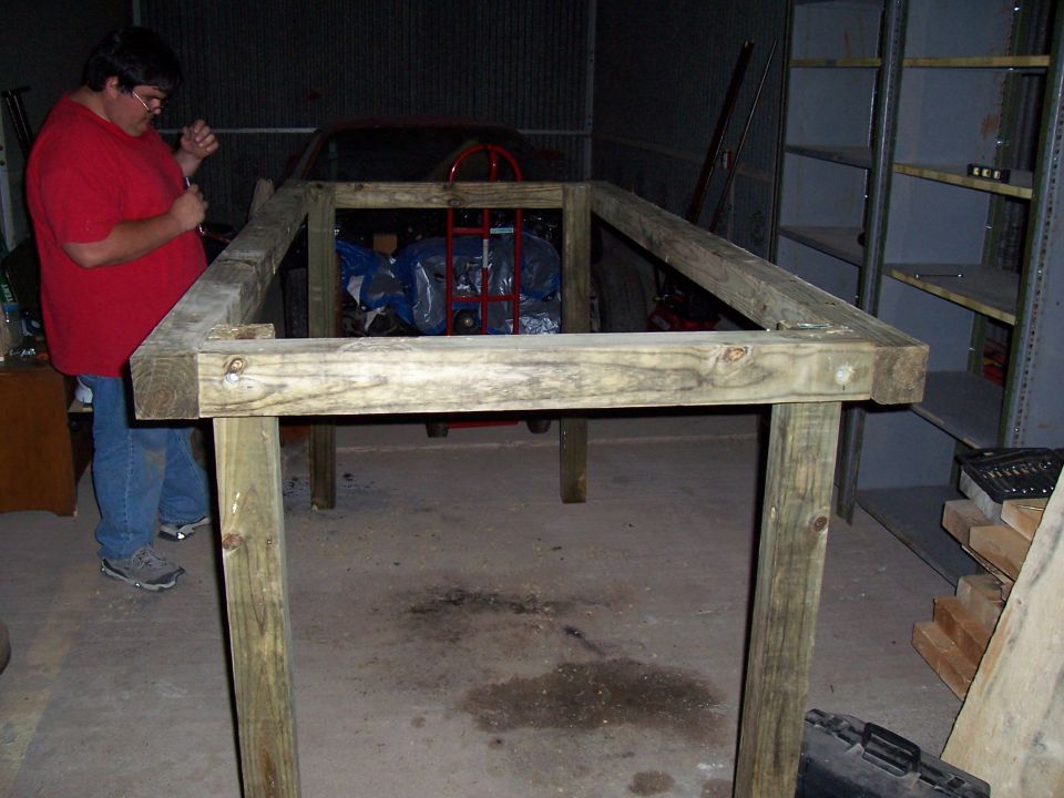 man in red shirt works on wooden table frame in garage, car partially visible behind him