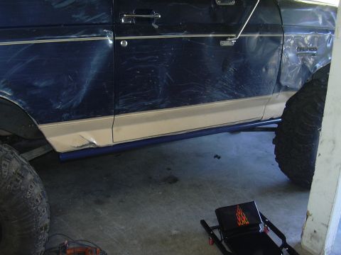 1991 Ford Bronco - Driver-side profile of a lifted dark blue Ford Bronco inside a garage, showing a newly installed blue-painted tube rock slider mounted alon…