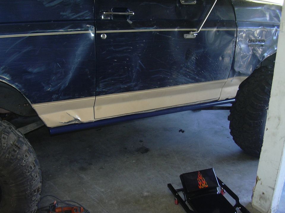 Blue tube rock slider mounted along the rocker of a lifted blue and tan Ford Bronco in a garage