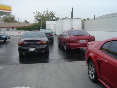 1991 Ford Taurus - Wet parking lot on an overcast day with a black third-generation Ford Taurus SHO and a red first-generation Taurus SHO parked rear-out besi…