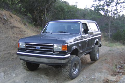 1989 Ford Bronco - A dark gray Ford Bronco is driven off-road on a dirt trail with trees in the background.