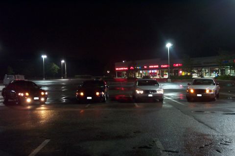 1999 Ford Crown Victoria - Four sedans lined up abreast with headlights on in an empty wet parking lot at night, a lit strip mall glowing in the background. The two o…