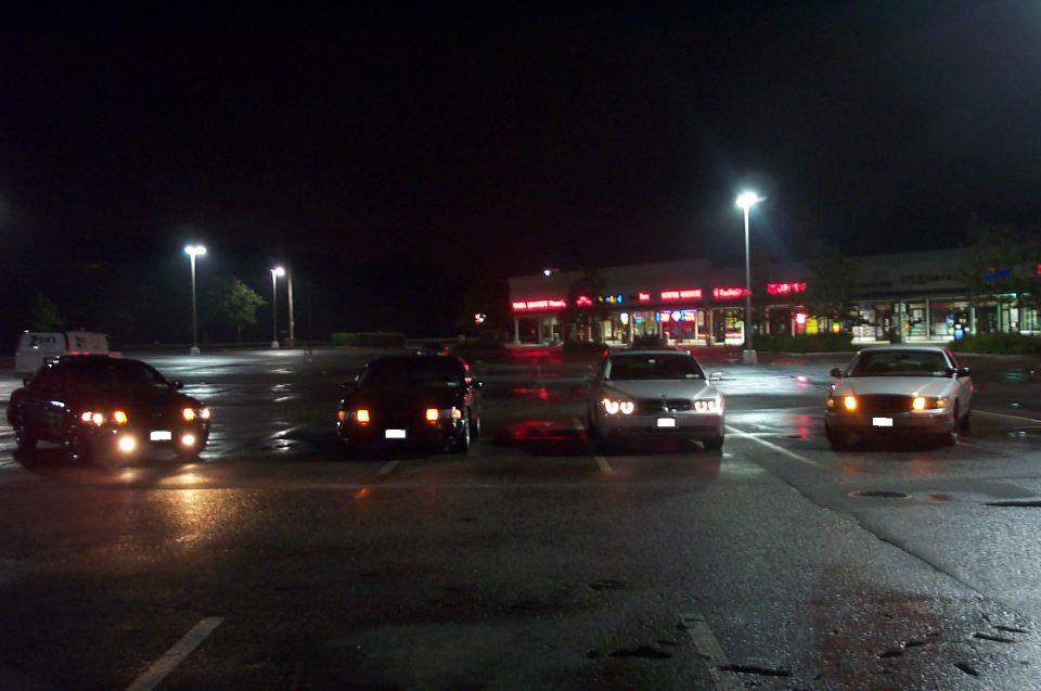 Four sedans lined up with headlights on in an empty wet parking lot at night